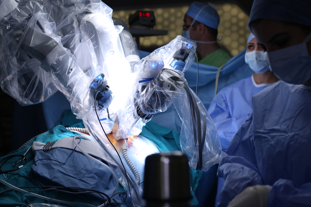 A imagem mostra braços robóticos realizando cirurgia em paciente deitado. Na foto, duas pessoas estão usando avental cirúrgico e parecem estar controlando o equipamento. Ilustração de tecnologia cirúrgica.