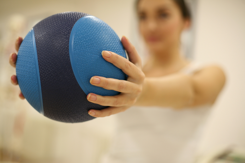Foto: Fisioterapia para reabilitação cardíaca/ cardiovascular.A foto mostra em primeiro plano uma bola azul utilizada para exercícios de fisioterapia. A bola está sendo segurada com as duas mãos por uma mulher que aparece em segundo plano, desfocada. Imagem ilustrativa de fisioterapia para reabilitação cardiovascular.